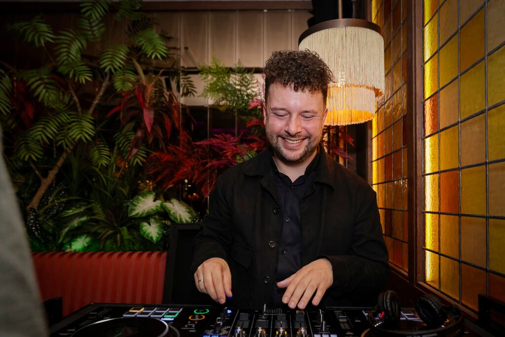 A DJ playing at a mixing desk in front of plants and amber glass windows