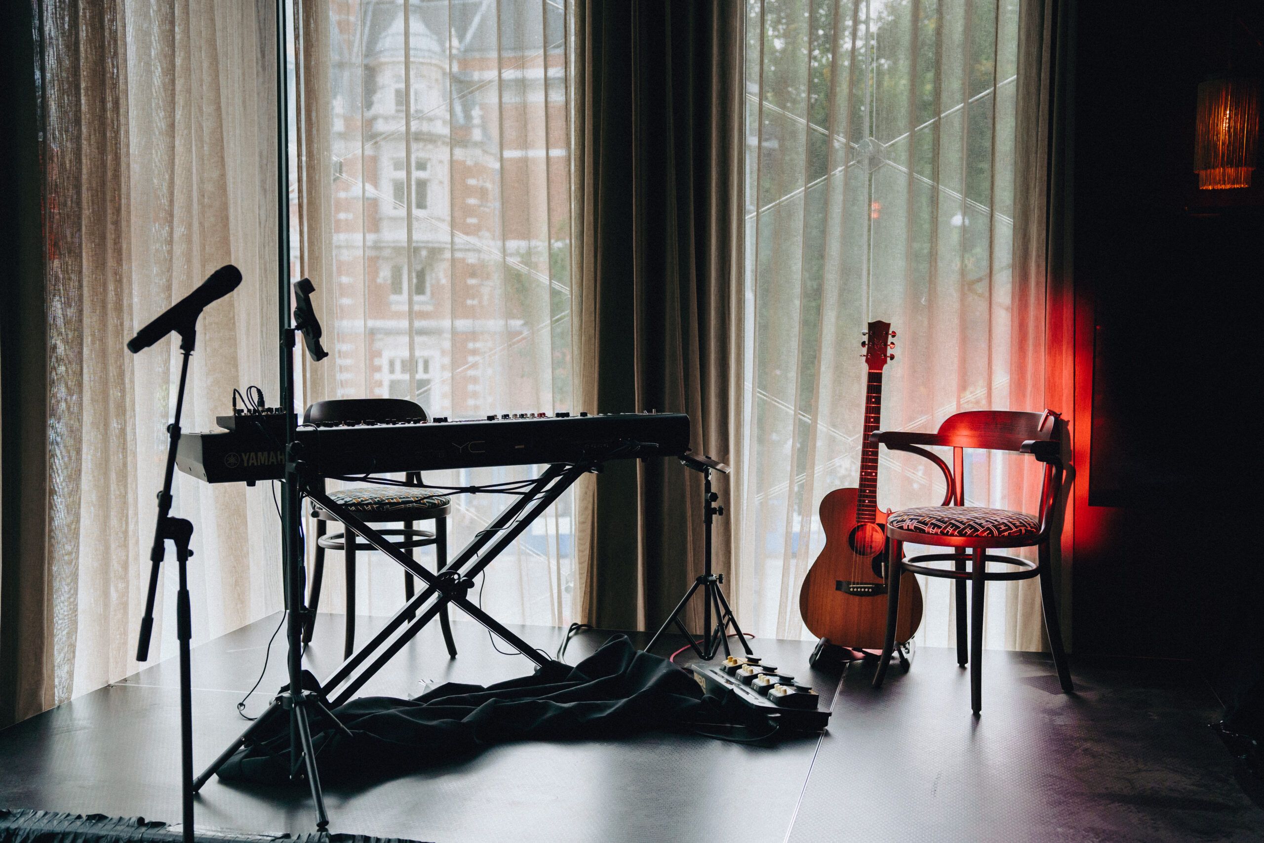 Small stage with a keyboard, microphone and acoustic guitar in front of tall curtained windows