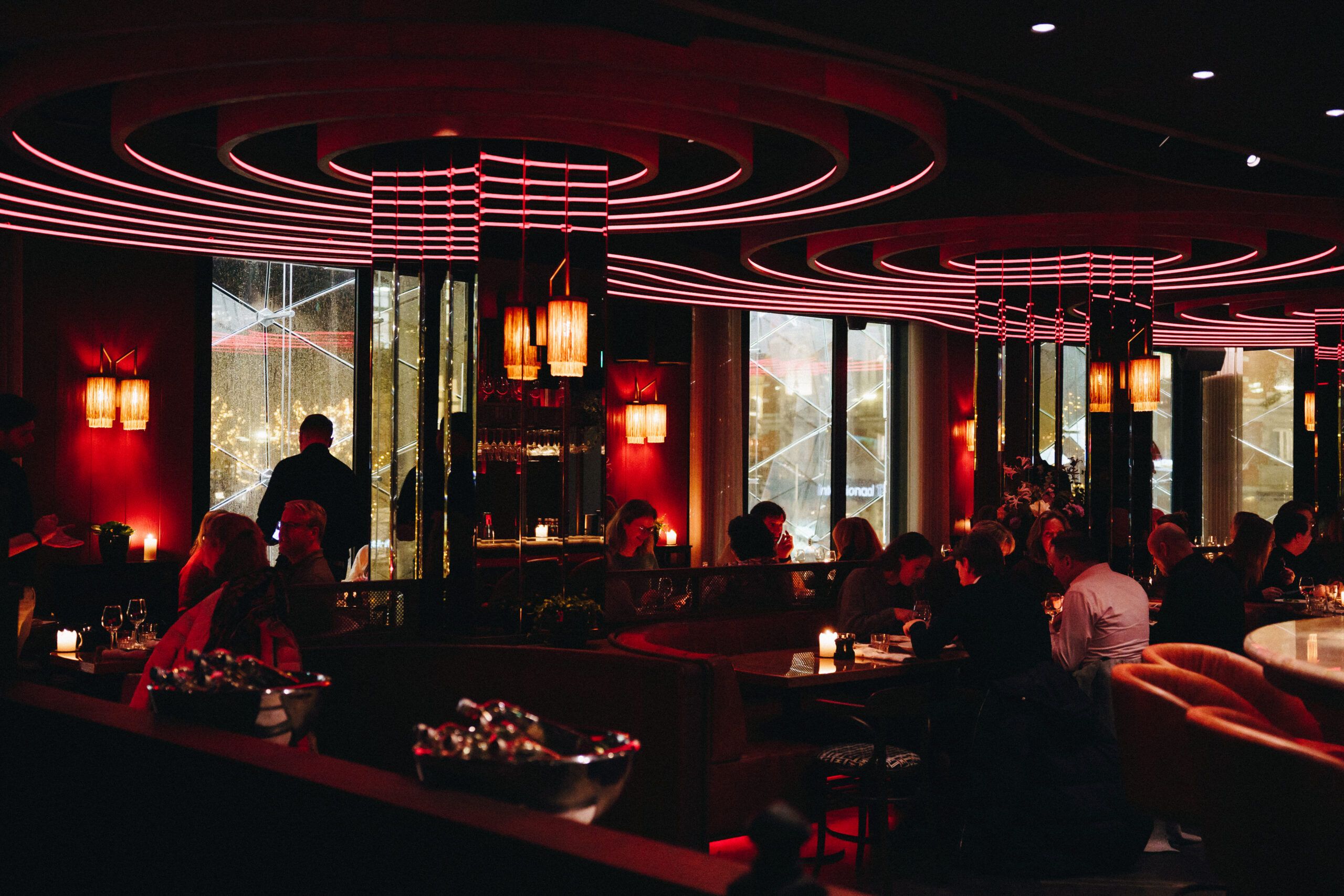 The dining room at night in red light under curved ceiling rings, with guests at the tables