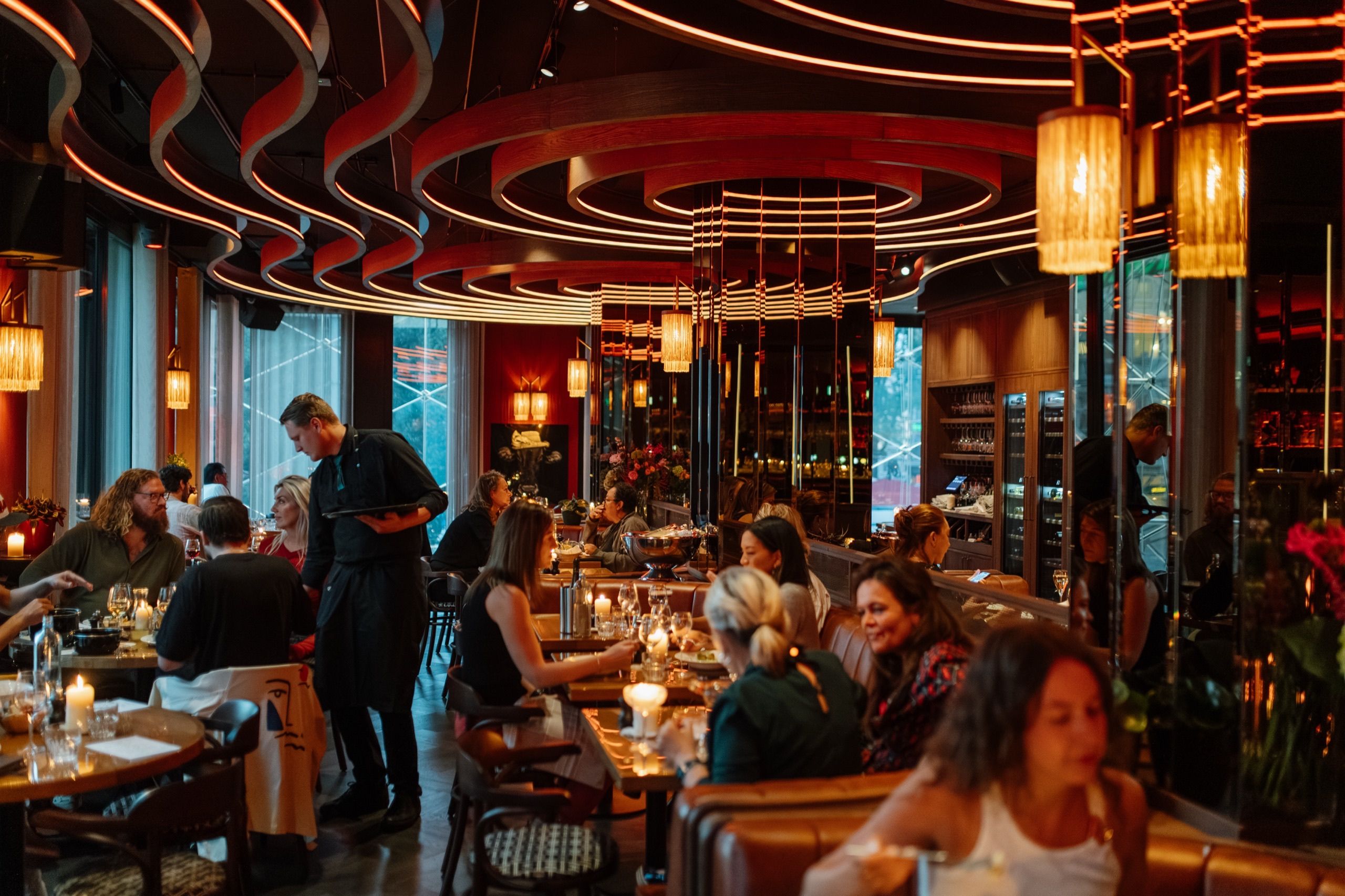 Een volle Bistro op de eerste verdieping in de avond: gasten aan tafels met kaarslicht onder het gebogen, verlichte plafond