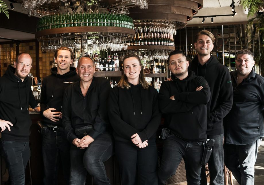 The Heineken Hoek team, seven people dressed in black, smiling at the Grand Café bar
