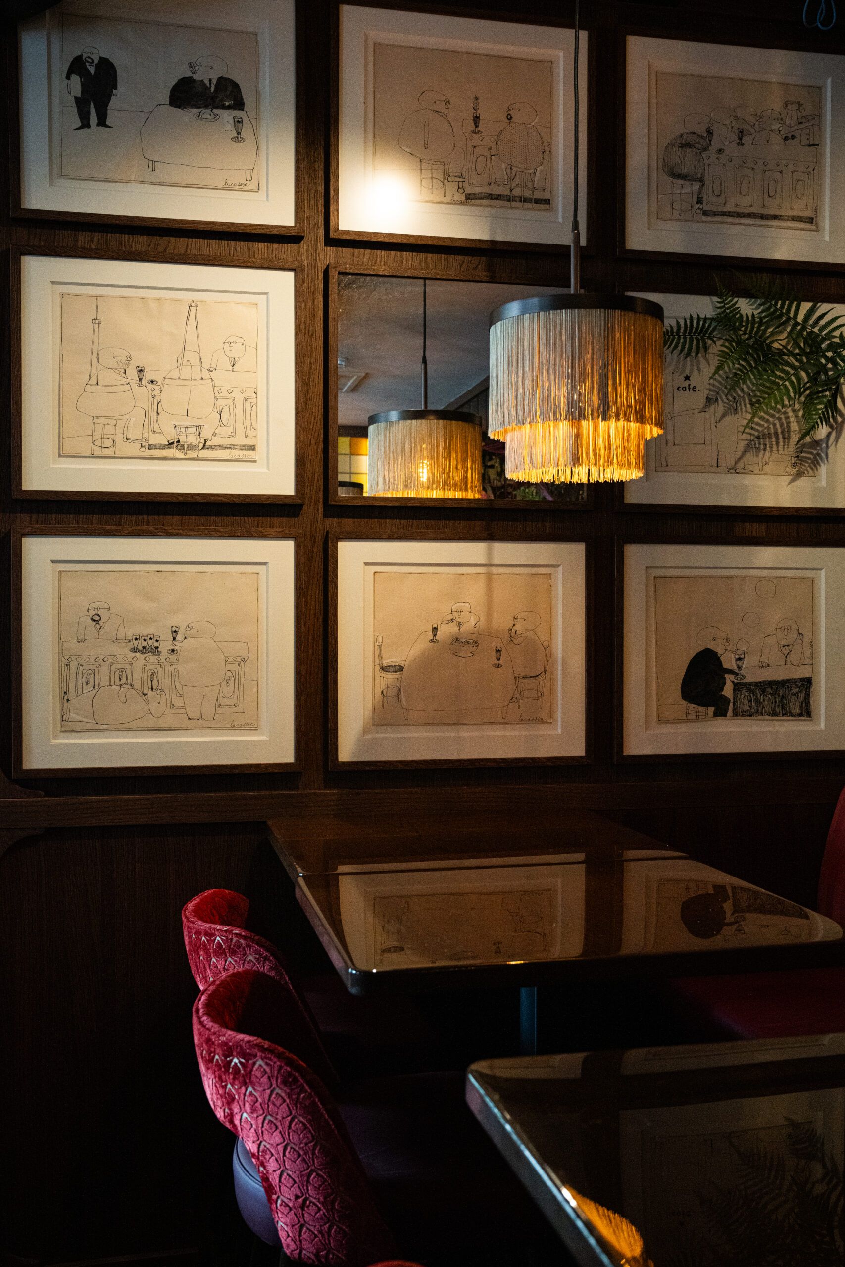 Framed drawings on a dark wood wall above a table with pink velvet chairs