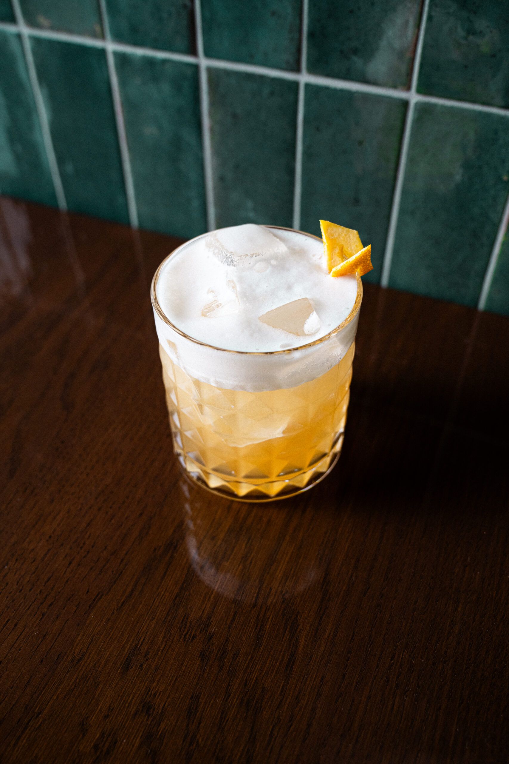 A frothy cocktail with an orange twist on a wooden table in front of green tiles