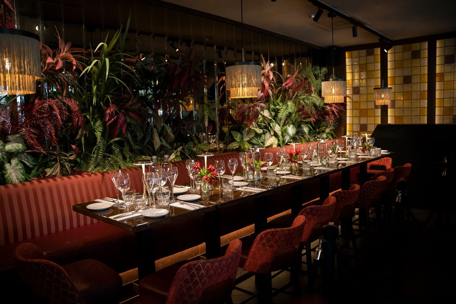 A long table set for a group dinner, with red velvet chairs, a striped banquette, plants and fringed pendant lamps